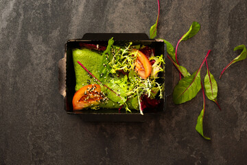 Spinach pancake with green lettuce, tomato and sesame salad in a black paper container on a dark background. Healthy food delivery service and daily ration concept.