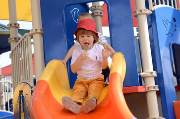 View from above at little boy eating green lollipop candy outdoors on playground, looking up to camera. Sweet tooth. A boy in a red panama. Portrait of a child.