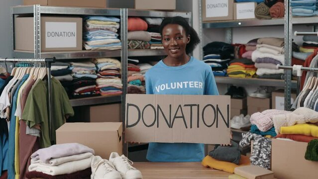 Portrait of energetic multiracial woman looking at the camera and smiling while holding banner with donation word in charity shop. Volunteer working in organization and sorting donated clothes