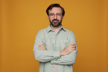 indoor portrait of handsome bearded male posing over orange background looking into camera with crossed hands on his chest