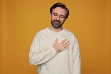 indoor portrait of handsome bearded male posing over orange background keeps his palm on heart, eye...