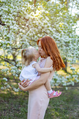Naklejka na ściany i meble A young woman kisses her young daughter in a beautiful flowering garden