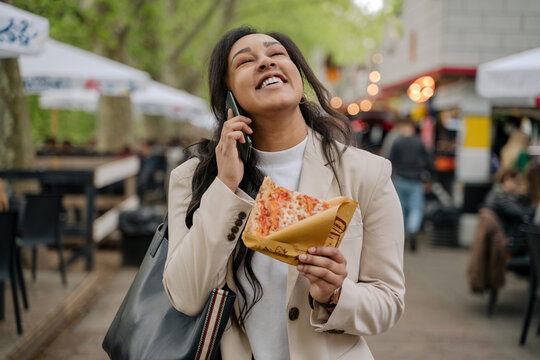 Fototapeta Authentic portrait of beautiful smiling woman talking on mobile phone holding slice of pizza walking on street market. Positive lifestyle concept 