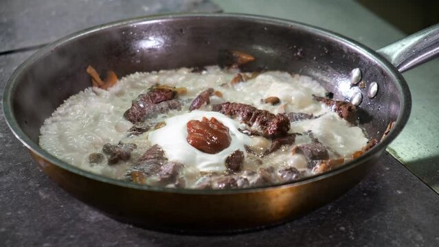 Chef in restaurant kitchen mixes ingredients in frying pan with wooden spoon mushrooms, onions, wild deer meat with cream and red sauce. Cooking for visitors. dish is fried and boils on stove