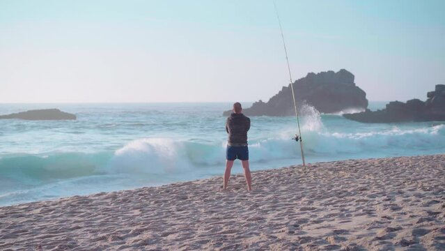Caucasian fisher man standing back to camera with fishing rod while fishing on the beach reels the line on the reel of fishing rod, pulls the fish out of the ocean, and raises it showing 