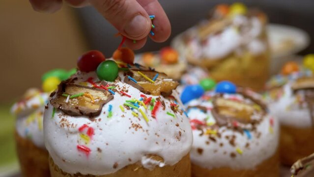 Female Hands Decorating Prepared Easter Cakes with Colorful Powder, Confetti. Yummy prepared yellow Easter cupcakes decorated with white icing, pieces of chocolate chips, nuts. Orthodox event Easter.