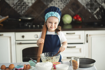 Cute little girl in apron and chef hat is flattening the dough using a rolling pin, looking at...