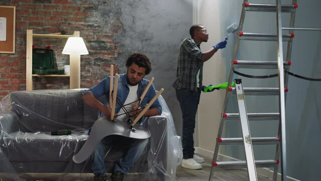 Black man paints wall guy assembles chair in living room