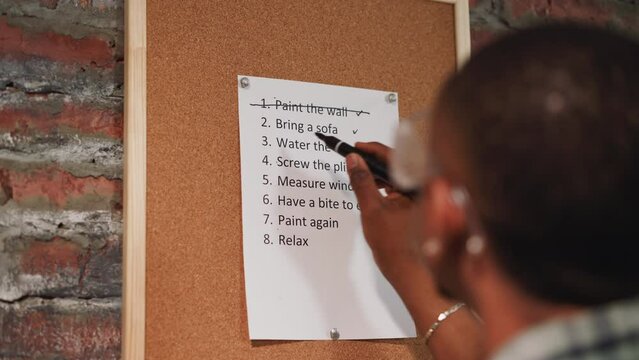 Black guy crosses out accomplished to-do list item on cork board near grunge wall closeup. African-American worker with plan of flat renovation process