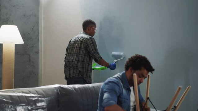 Black man applies paint on wall friend fixes chair in room