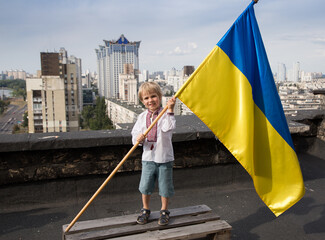 boy in an embroidered shirt stands on the roof and holds a large Ukrainian flag against the backdrop of the cityscape of the city of Kyiv. National symbol of freedom and independence. Stop the war