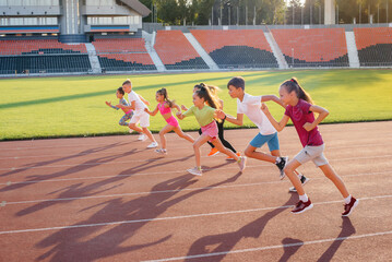 A large group of children, boys and girls, run and play sports at the stadium during sunset. A...