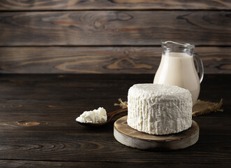 Fresh milk curd lies on a round wooden plank. A jug of milk in the background. Wooden spoon with cottage cheese on the table. Light wooden background, space for text