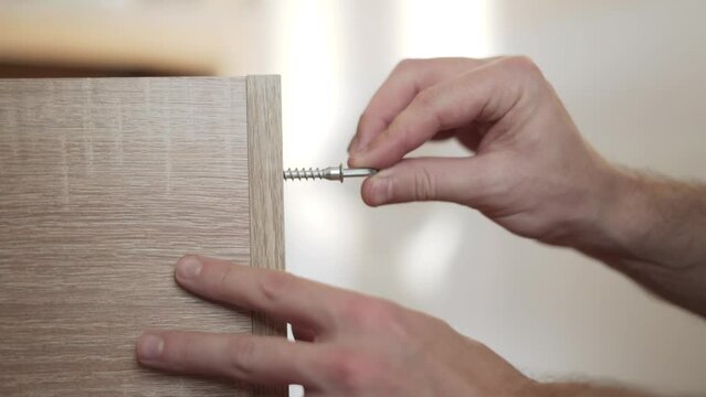 Male caucasian hands manually twist screw with metal hex key, close-up. Euro screw fastens the details of the chipboard cabinet at butt. The work of a furniture assembler.