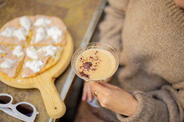 Woman holding wooden board with small pizza and glass of shakerato drink at outdoor restaurant, close-up, cropped view focused on cocktail. Concept of italian cuisine while travel