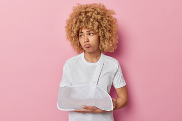 Sad woman has broken arm in splint has unhappy mood remembers personal accident looks displeased away wears casual white t shirt isolated over pink backgground. Female injured after falling.