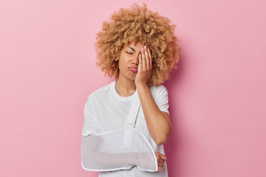 Displeased doleful curly haired woman makes face palm feels frustrated makes face palm has broken arm in splint as result of work trauma dressed in casual t shirt isolated over pink background.