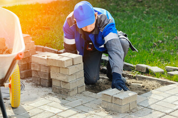 Bricklayer in work clothes sits on sidewalk and lays out paving slabs. Sight of working man in open...