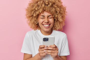 Joyful curly haired woman types text messages surfs internet and laughs sincerely reads funny content in internet giggles positively dressed in white casual t shirt isolated over pink background.