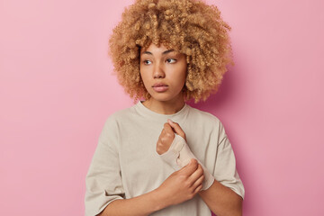 Photo of serious thoughtful curly woman looks away wears elastic bandage on broken arm has painful treatment dressed in casual t shirt isolated over pink background. Female model after accident