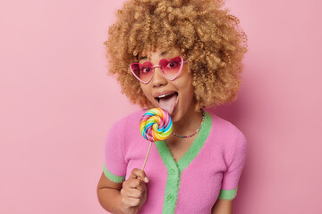 Cheerful curly haired woman licks delcious caramel multicolored round candy being sweet addicted wears trendy pink sunglasses casual shirt isolated over pink background. Sugar nutrition concept