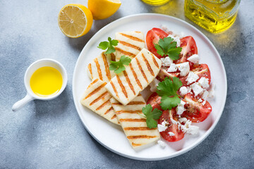 Grilled halloumi cheese with red tomatoes and feta on a white plate, high angle view on a light-blue stone background, horizontal shot