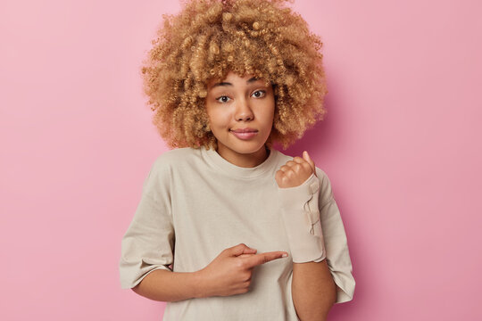 Young curly haired European woman shows broken arm after accident wears elastic bandage dressed in casual t shirt isolated over pink studio background. Insurance accident and health care concept.