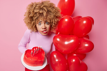 Irritated surprised woman stares at camera purses lips with annoyance has leaked makeup holds delicious heart shaped heart and bunch of red balloons isolated over pink background. Festive occasion