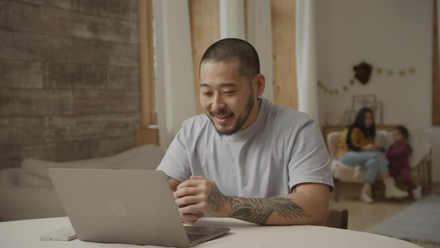 Young adult father talking to his relatives on a video call with his family playing in the room