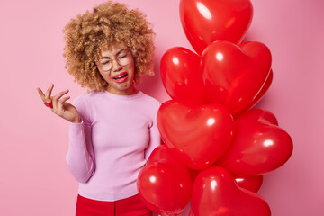Dejected frustrated curly haired woman cries from despair looks doleful has spoiled makeup dressed in casual clothes celebrates lovers day alone poses near bunch of red balloons poses indoor