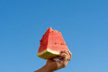 Male hand holding a slice of watermelon against a blue sky.