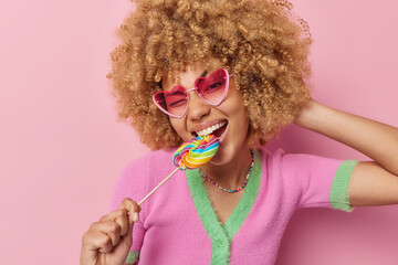 Happy carefree curly haired woman cannot live without sweets bites multicolred candy wears heart...