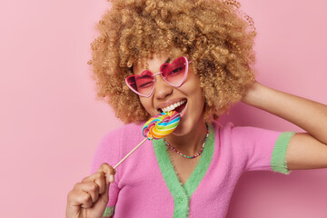 Happy carefree curly haired woman cannot live without sweets bites multicolred candy wears heart...
