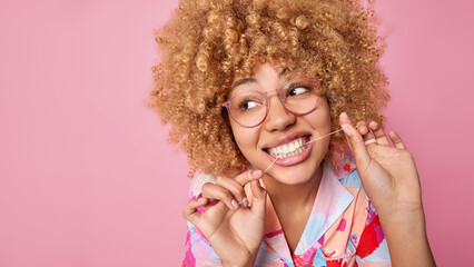Close up shot of curly haired woman uss dental floss takes care of oral hygiene looks away wears...