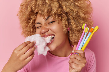 Horizontal shot of curly haired young woman bites appetizing candy floss harmful for teeth holds...