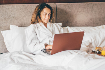 Woman eating breakfast and working at laptop in the hotel room.