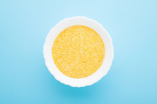 White bowl with dry yellow cornmeal porridge on light blue table background. Pastel color. Closeup. Healthy food. Top down view.