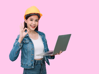 Portrait of cheerful asian young woman engineer in jeans dress and wearing yellow safety hardhat, holding laptop for work isolated on pink background