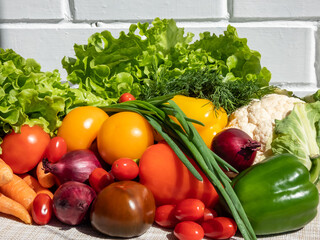 A pile of fresh, healthy and colorful vegetables in bright sunlight with white brick wall in background. Tomatoes, onions, carrots, peppers, cauliflower, salad, dill and greens