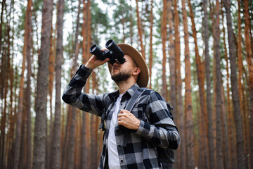 Young Man in a hat with a backpack and binoculars in a pine forest. Hike in the mountains or forest