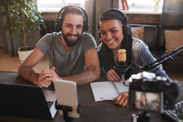 Horizontal high angle shot of two happy multi-ethnic influencers sitting at desk in loft room smiling at smartphone camera