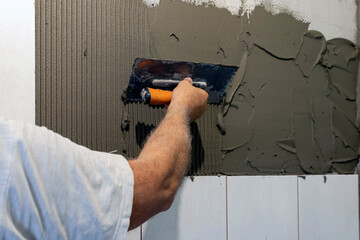 Repair in the apartment: laying tiles on the walls. / A worker coats the wall with glue with a...