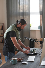 Vertical long shot of young bearded man adjusting audio mixer setting sound in home studio before starting shooting content for blog