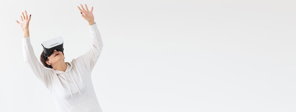 Unidentified middle-aged woman in a light sweater plays a 3D game with glasses of virtual reality on banner white background with copy space. High technology concept for older people.