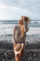 Portrait of a beautiful blonde-haired young woman on the beach with a beige sweater. She is holding a straw hat