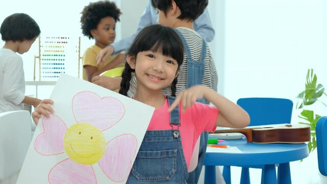 Happy little girl show the picture after to paint color book in classroom,kindergarten education school.Multi-ethnic preschool teacher and students in classroom.Kindergarten and study concept.