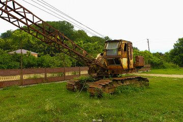An old rusty crawler crane. Standing on the green grass near the fence.