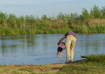 father holding a small child in his arms trying to wash her feet from the sand in the water in the lake around the green grass clear water blue sky shining sun summer vacation