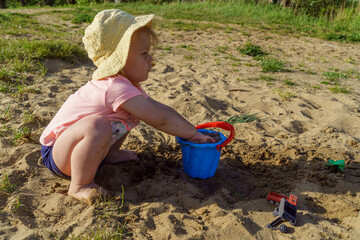 little baby playing with a blue plastic bucket filled with water on a background of sand, a child with bare feet pours water from a bucket learns something new fun childhood in the summer on vacatio