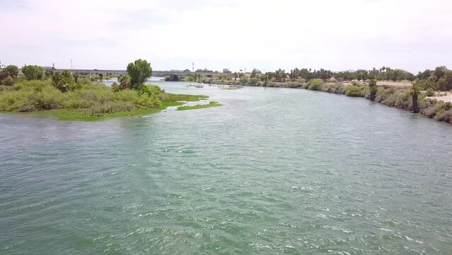 aerial footage of the silky green waters of the Colorado River with people in boats on the water surrounded by lush green palm trees and grass with blue sky at Quechan Park in Blythe California USA